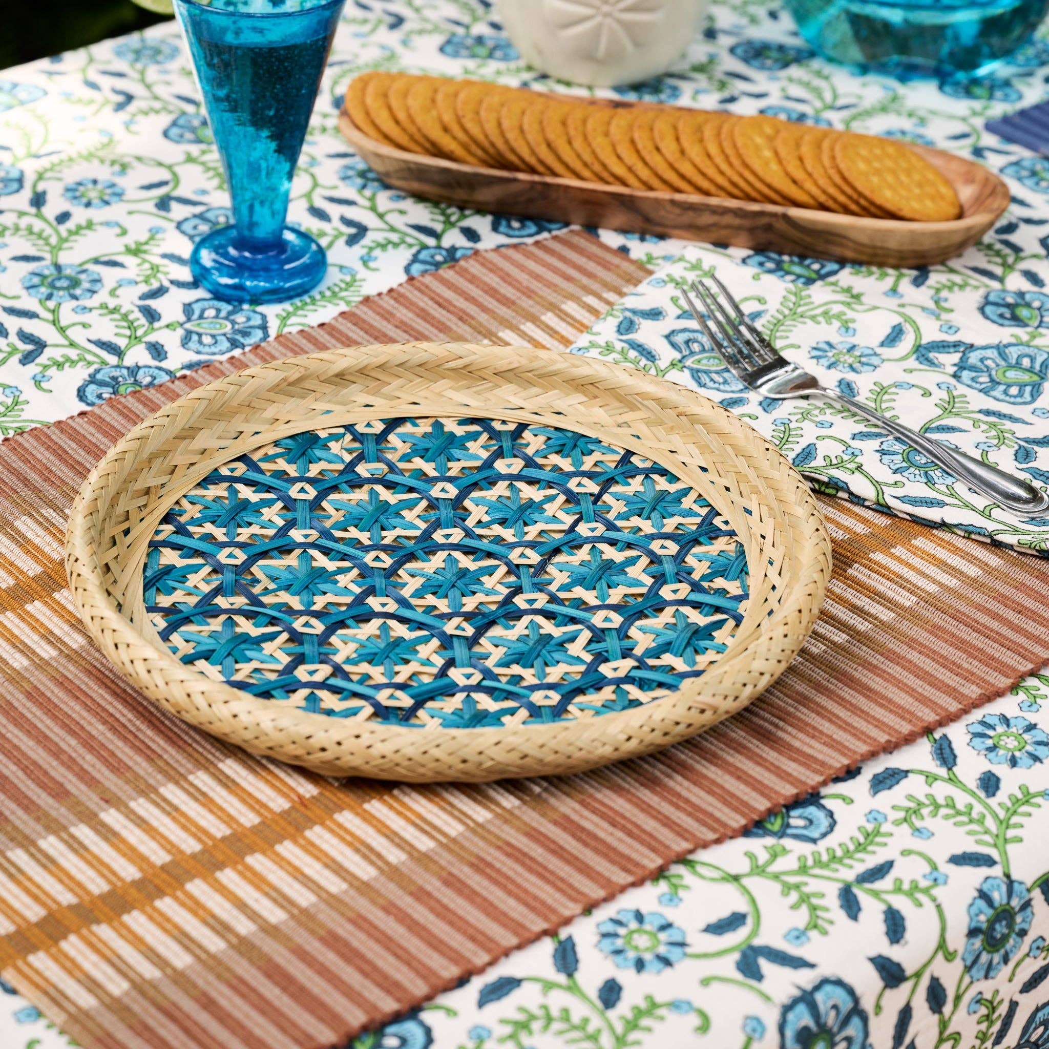 Lifestyle — placemat on floral tablecloth with woven tray and crackers: "Brown striped handloomed cotton placemat on a floral tablecloth, styled with a woven rattan tray, crackers, and a blue glass"