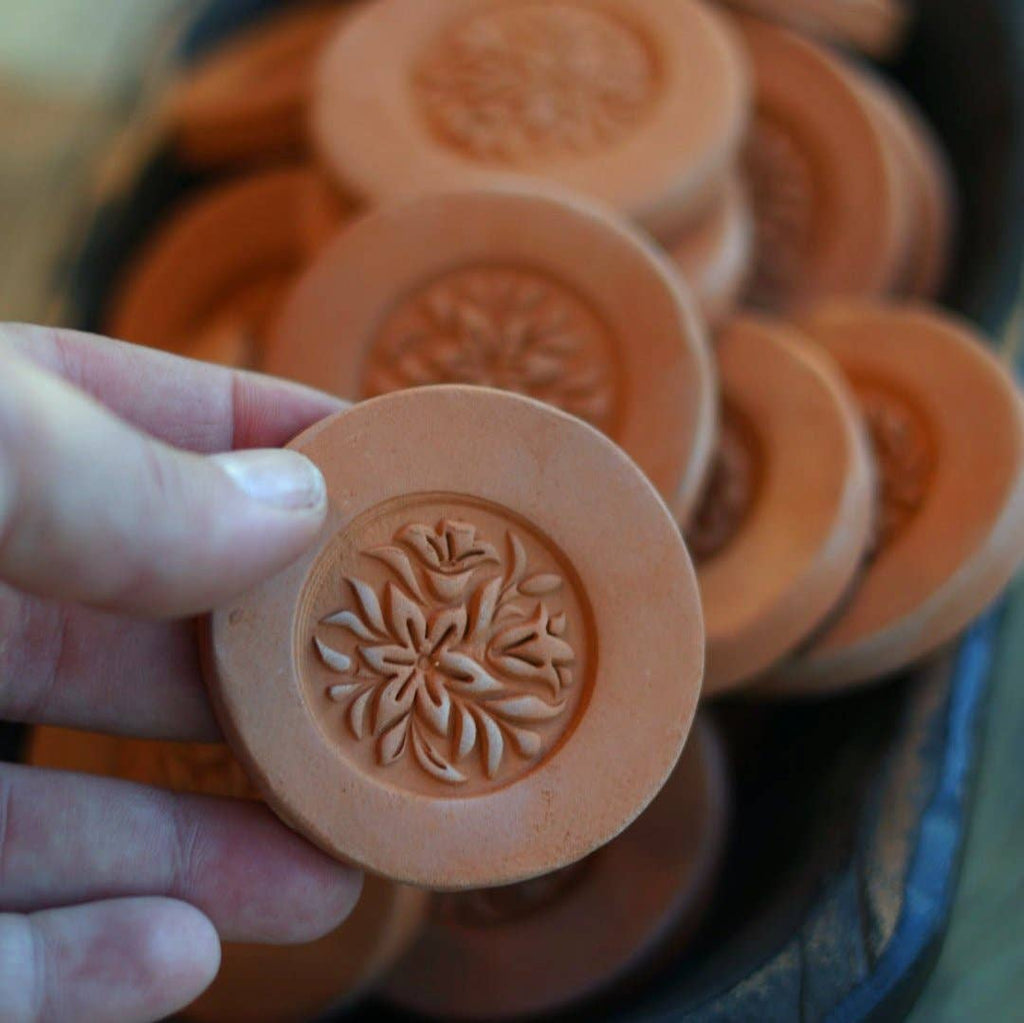 "Hand holding a single Gravesco handmade terracotta brown sugar keeper showing detailed floral medallion carving, with more in background"