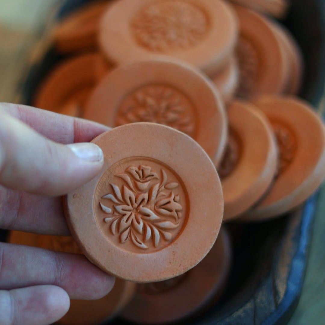 "Hand holding a single Gravesco handmade terracotta brown sugar keeper showing detailed floral medallion carving, with more in background"
