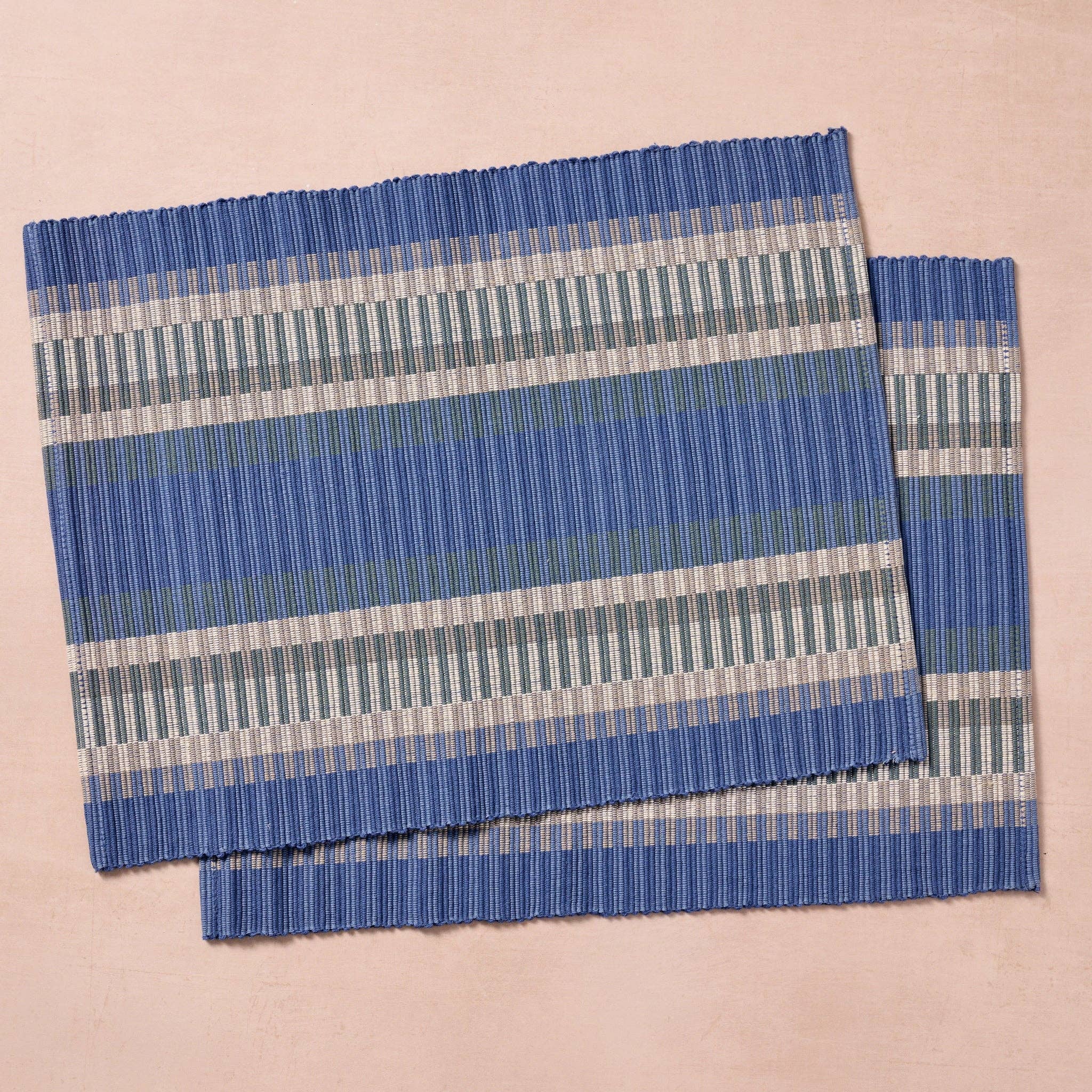 Two placemats laid flat (pink bg): "Set of two blue, gray, and white striped handloomed cotton placemats, handcrafted in Nepal by Ten Thousand Villages artisans