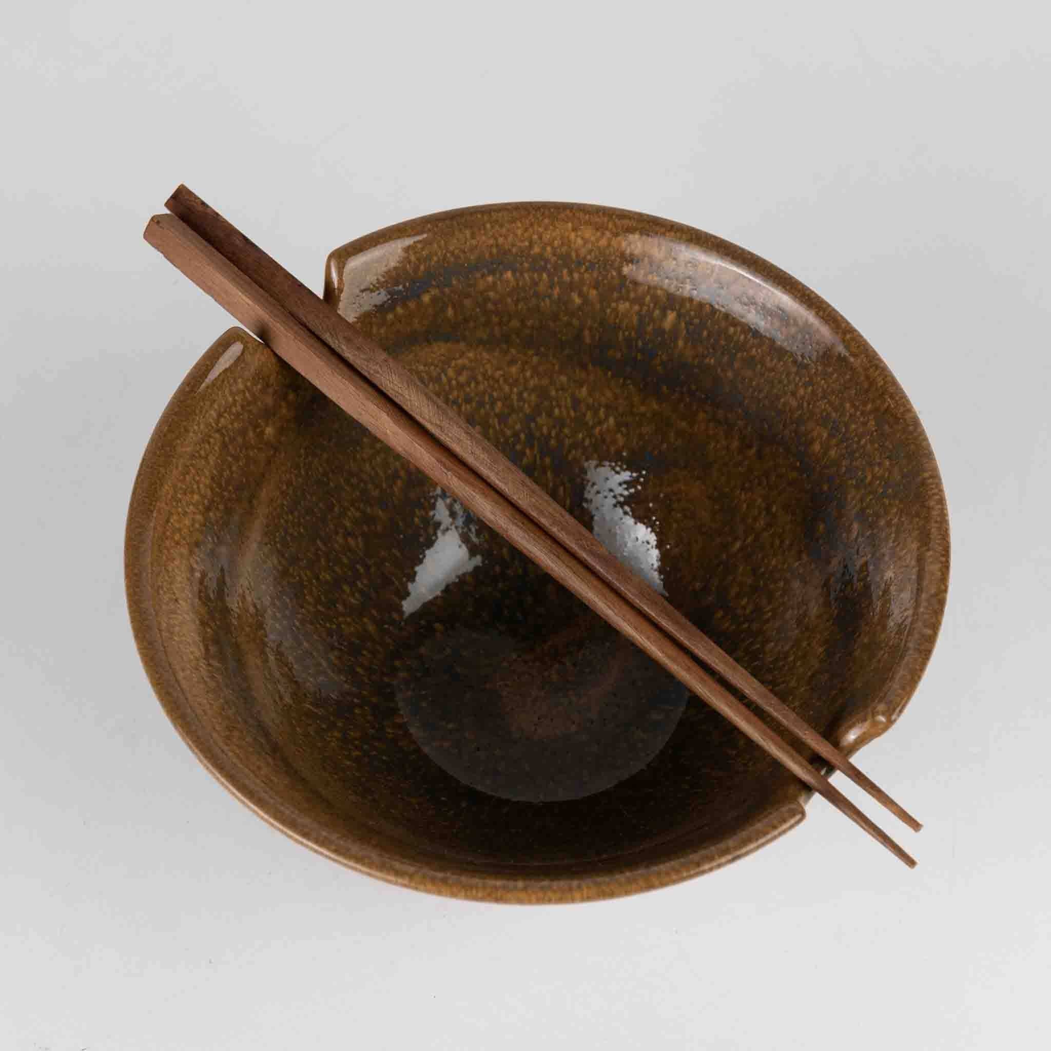 Overhead view of bowl with chopsticks: "Overhead view of handmade terracotta ceramic ramen bowl with Saj wood chopsticks resting across the notched brim"