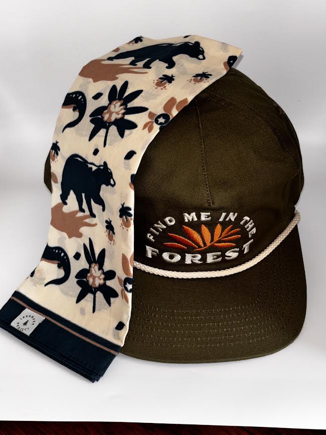 An olive green baseball cap with embroidered text reading 'FIND ME IN THE FOREST' and a matching patterned scarf draped over the brim. The scarf has a beige base with black animal silhouettes, brown shapes, and small orange floral accents.