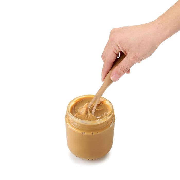 Hand using the HIC Kitchen nut butter mixer to stir natural peanut butter in a glass jar, demonstrating the open paddle blending separated oils back into the butter.