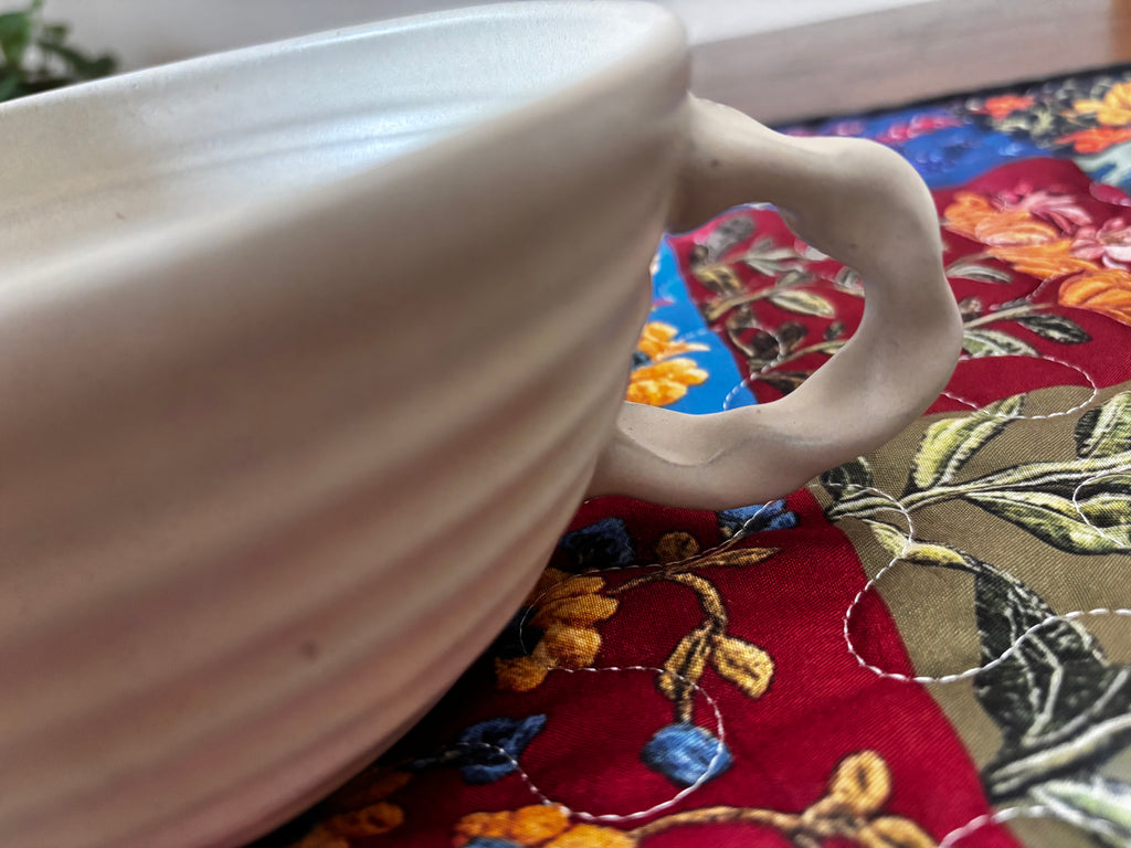 "Close-up of handmade ceramic mixing bowl handle — organic sculpted cream clay handle on colorful quilted runner"