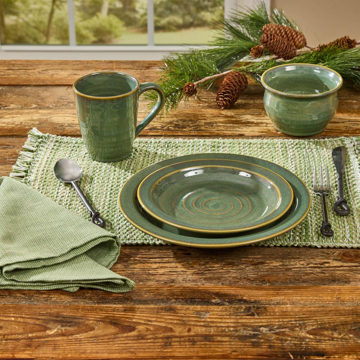 Park Designs Sinclair Caprese cereal bowl and mug on a farmhouse wood table with green woven runner, pine branches, pine cones, and sage green napkin"
