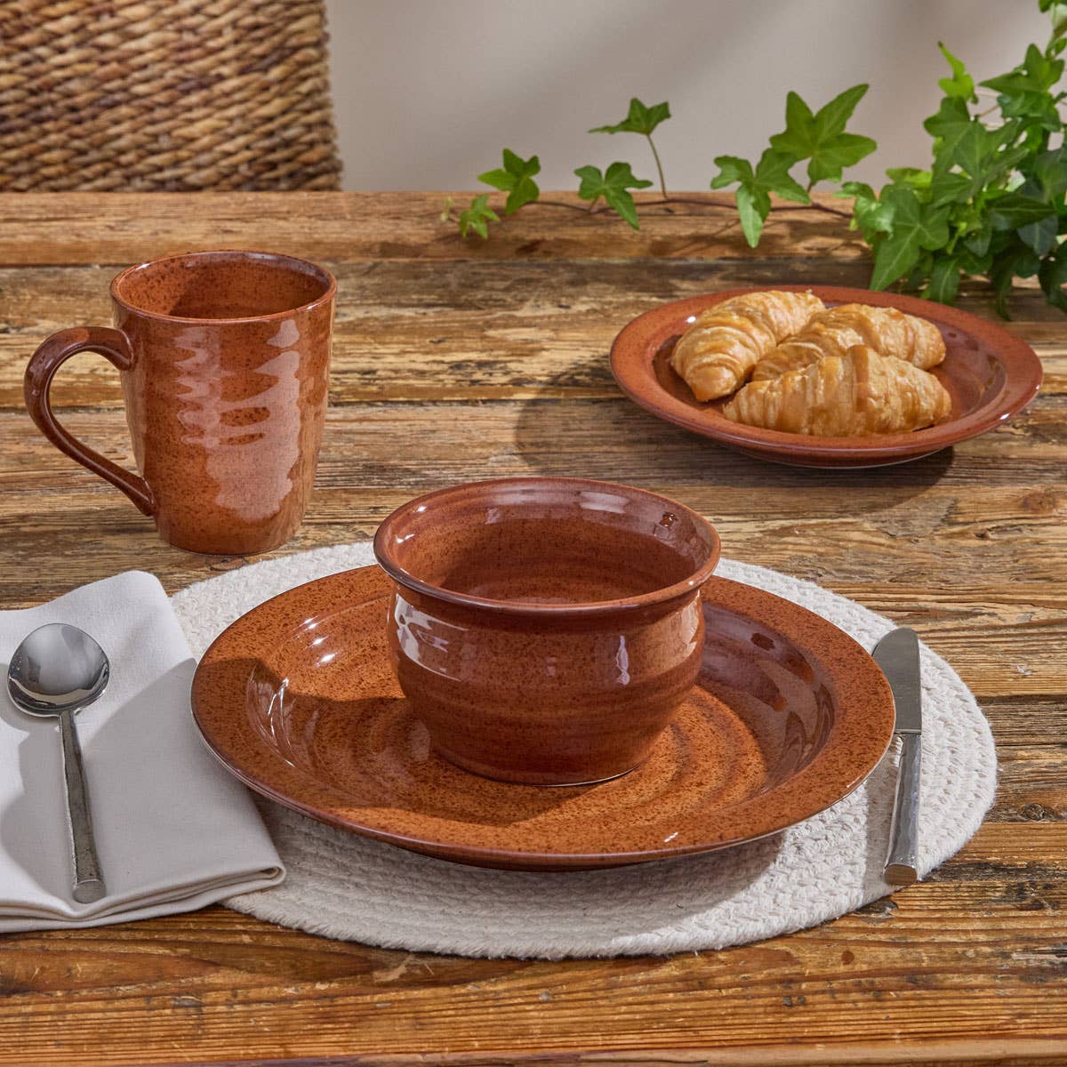 Park Designs Sinclair Tomato Soup cereal bowl and salad plate on a white woven placemat with matching mug, silver cutlery, and fresh croissants on a farmhouse wood table"