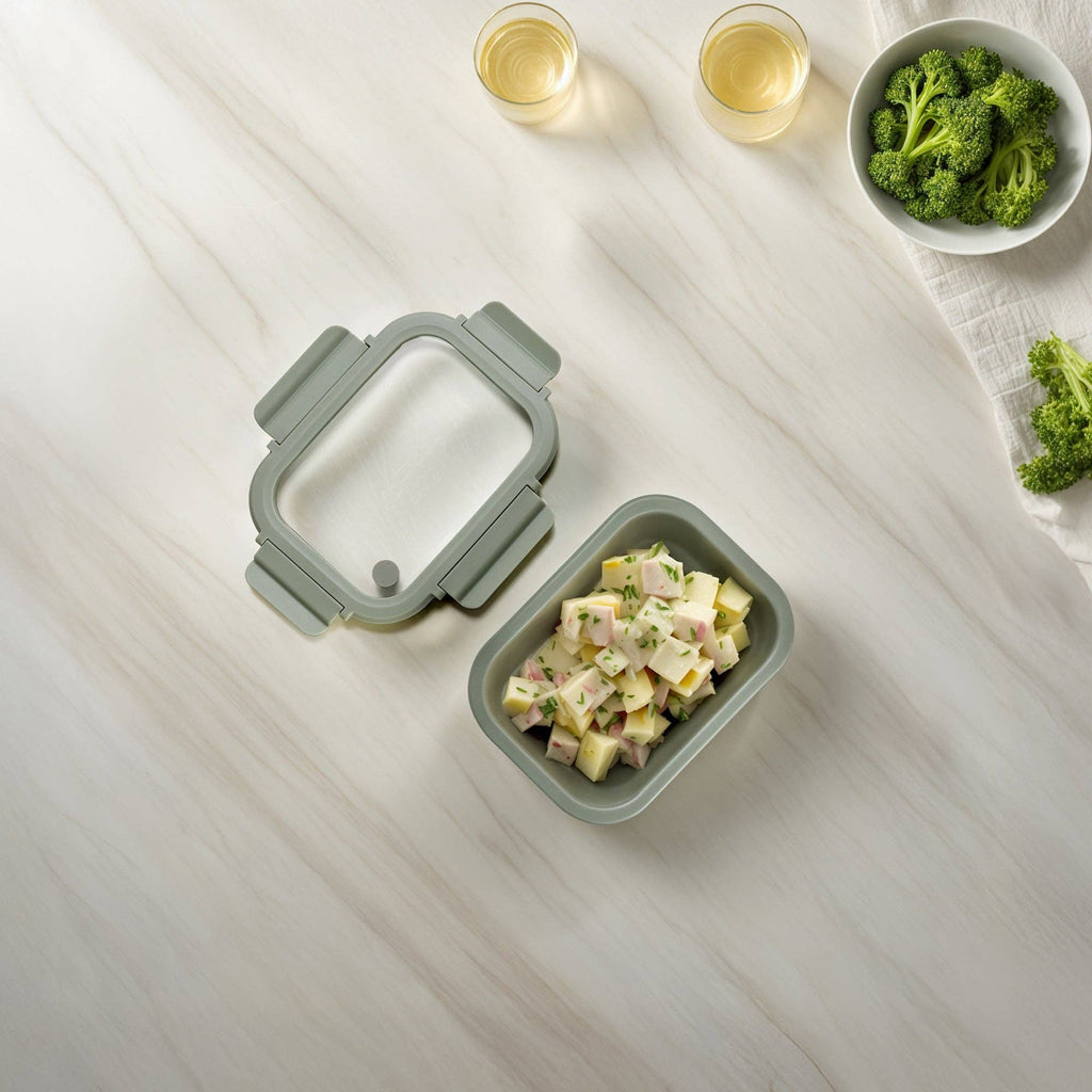 Green food container and lid with salad on a light wooden surface with broccoli and wine glasses.