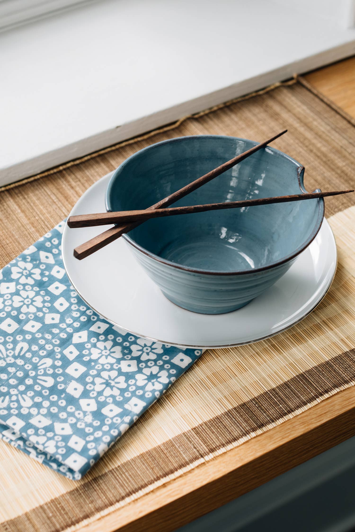Lifestyle shot on wooden surface with blue napkin: "Blue stoneware ramen bowl and chopsticks set styled on a wooden surface with a blue floral napkin"