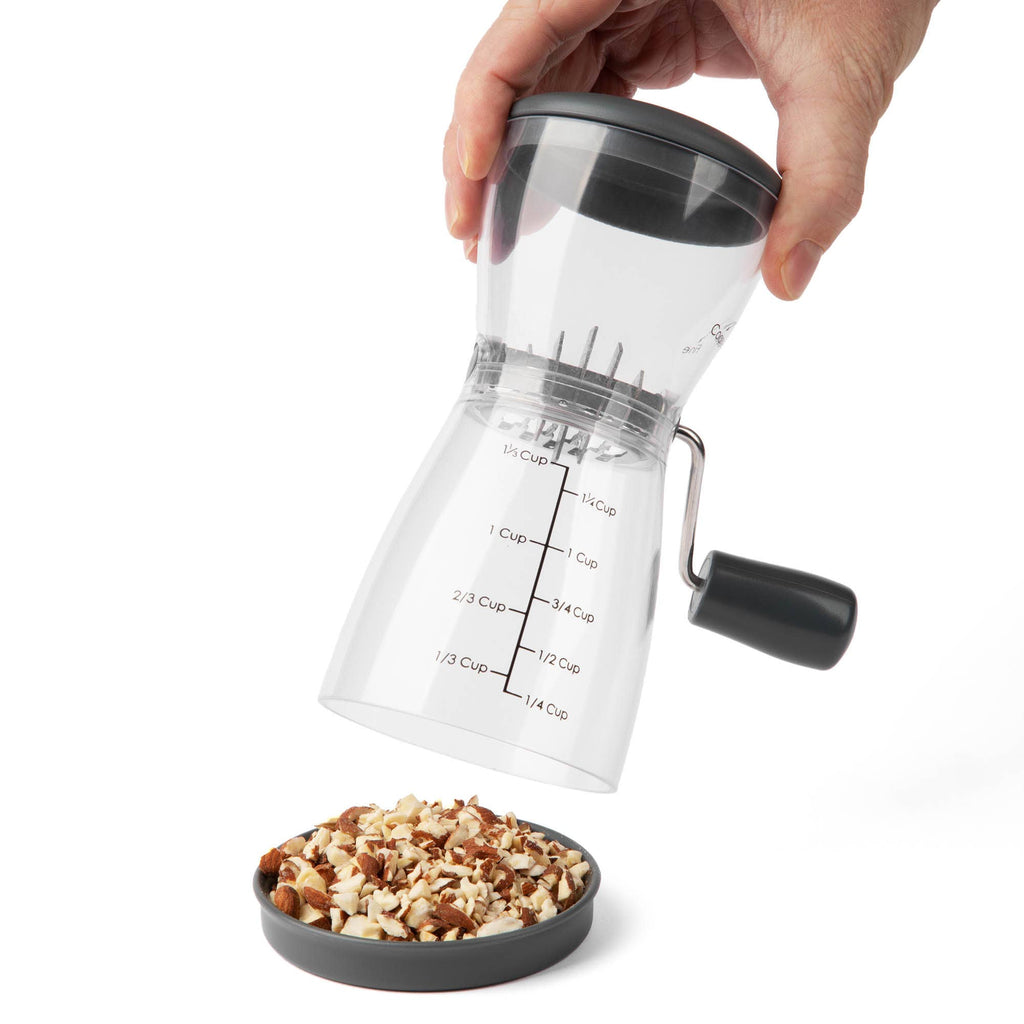 Hand lifting the lid of the Progressive nut chopper over a small gray dish filled with coarsely chopped mixed nuts, demonstrating the removable top for easy emptying.
