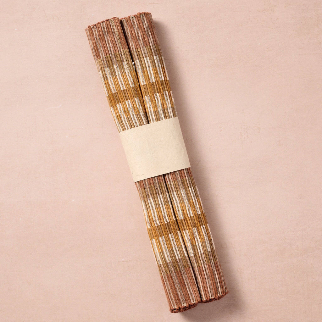 Single placemat rolled and banded (blush bg): "Single handloomed brown, tan, and gold striped cotton placemat rolled and wrapped with a paper band, handcrafted in Nepal"