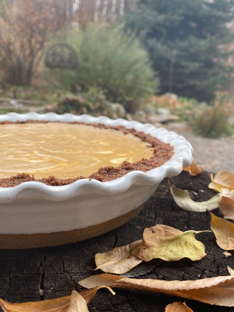 White speckled stoneware pie dish with ruffled edge holding a golden custard pie, styled outdoors on a wood surface surrounded by autumn leaves