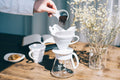 Hand pouring coffee grounds into a Hario V60 white ceramic pour over dripper with paper filter, styled on a wood table with dried flowers and a mug