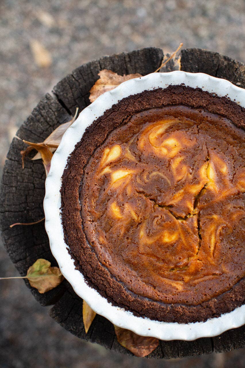 Overhead view of a dark chocolate swirl cheesecake in a white speckled stoneware pie dish with ruffled edge, resting on a tree stump surrounded by fallen autumn leaves