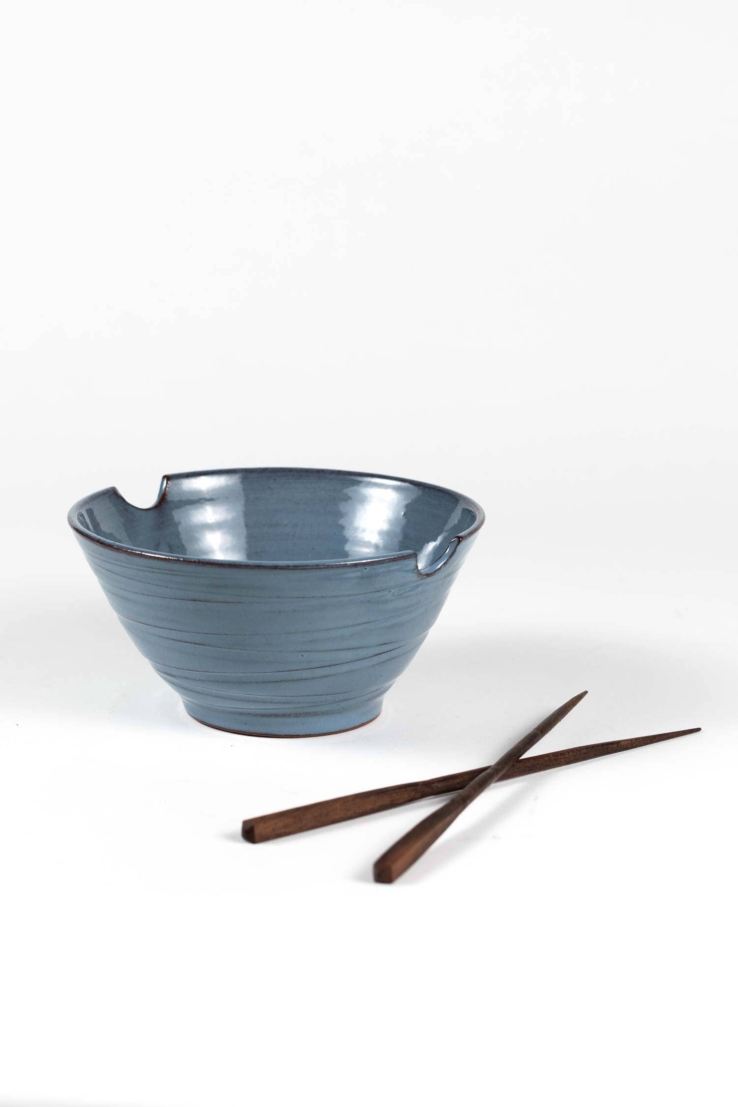 Bowl and chopsticks side by side (white bg): "Blue stoneware ramen bowl with chopstick rest grooves and Saj wood chopsticks, part of the Indigo Night collection by Ten Thousand Villages"