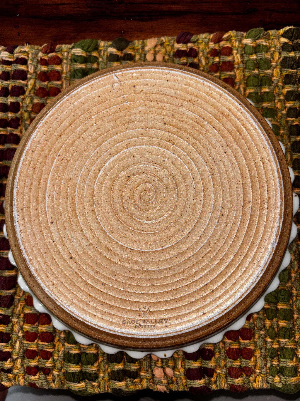 Unglazed natural clay base of a 6-inch Sage Valley Pottery deep dish pie plate showing hand-thrown spiral rings and stamped maker's mark on a woven textile