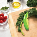 Green vegetable peeler on a wooden cutting board with kale and tomatoes in the background.