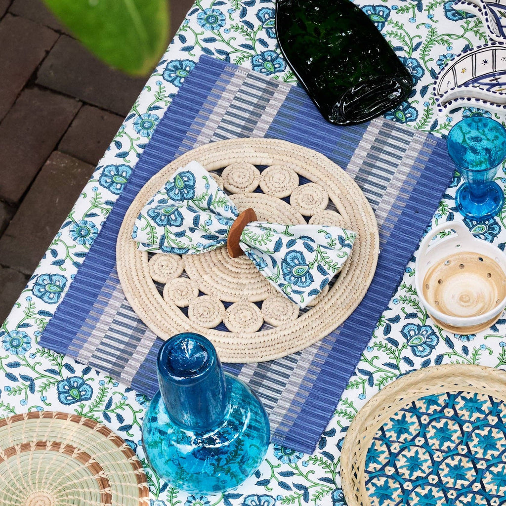 Outdoor table lifestyle shot: "Blue striped handloomed cotton placemat styled on an outdoor table with a floral tablecloth, woven charger, blue glassware, and a folded napkin"