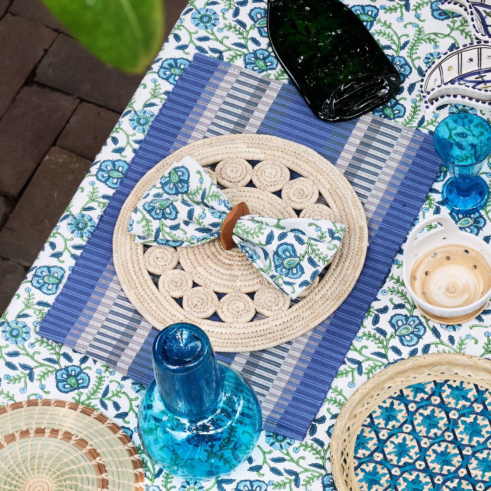 Outdoor table lifestyle shot: "Blue striped handloomed cotton placemat styled on an outdoor table with a floral tablecloth, woven charger, blue glassware, and a folded napkin"