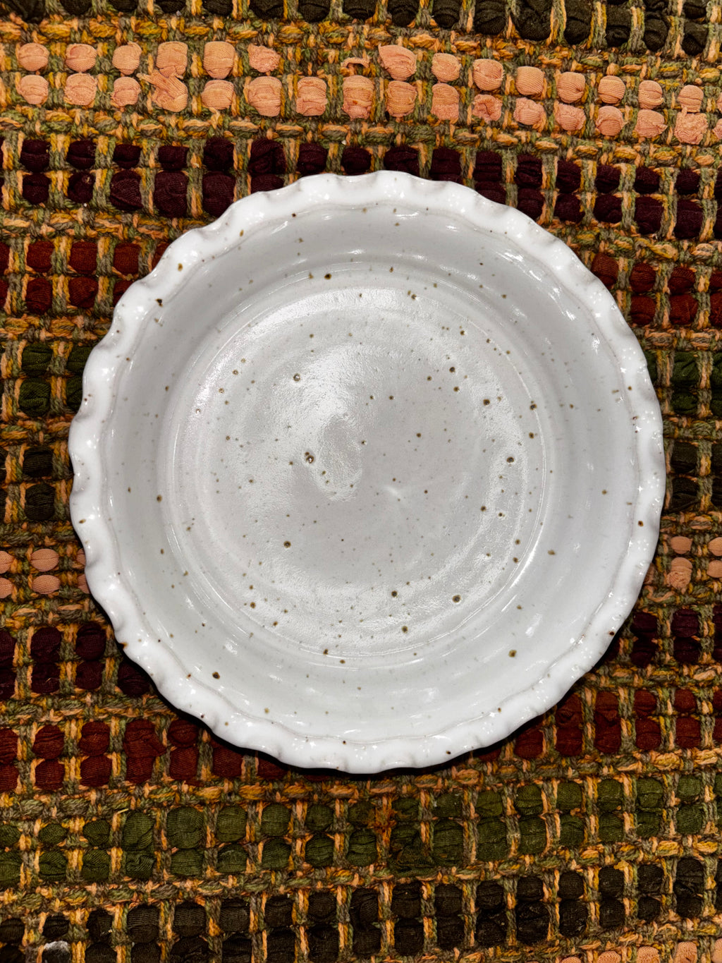 Overhead view of a 6-inch hand-thrown white speckled stoneware deep dish pie plate with ruffled scalloped edge on a woven multicolor textile