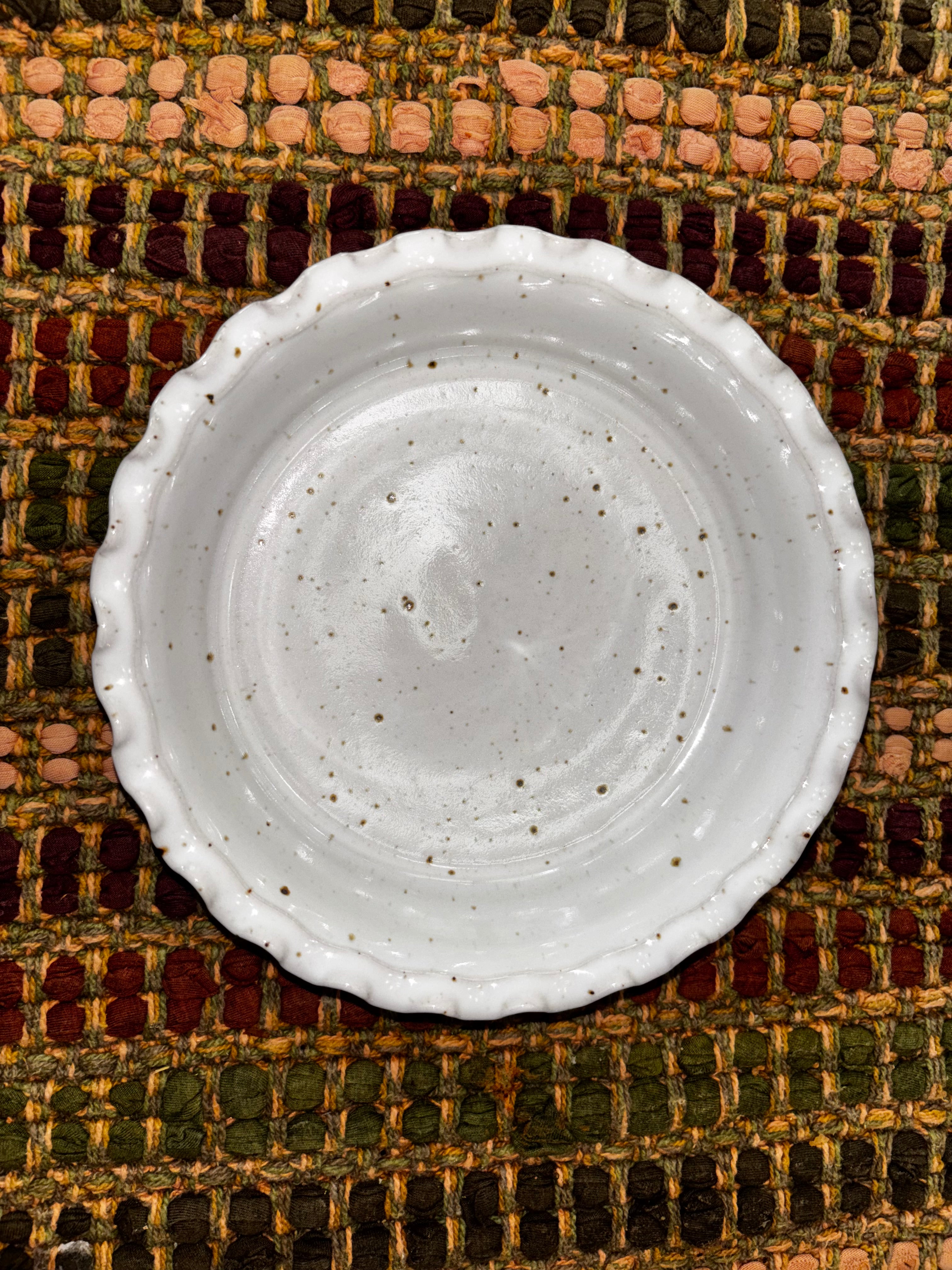 Overhead view of a 6-inch hand-thrown white speckled stoneware deep dish pie plate with ruffled scalloped edge on a woven multicolor textile