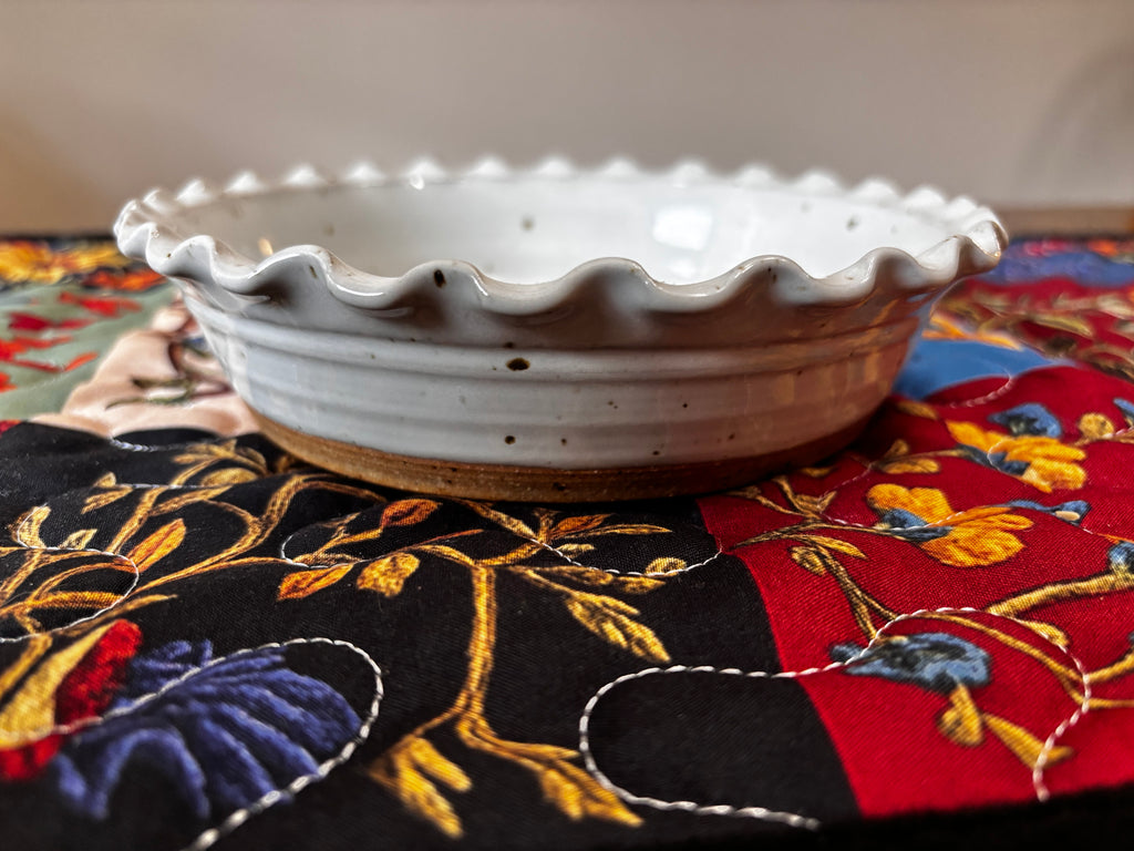 6" side profile on floral quilt) Side profile of a 6-inch hand-thrown white speckled stoneware deep dish pie plate with ruffled edge and natural clay base, resting on a colorful floral