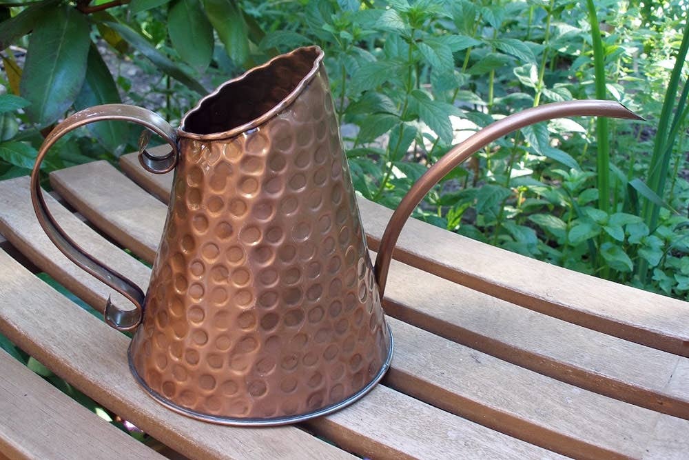 Hammered copper watering can with scrolled handle and long curved spout displayed on a wooden garden bench with green foliage in the background