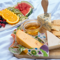 Bee's Wrap Garden Party beeswax wrap in colorful fruit print loosely wrapped around a wedge of cheese on a wood charcuterie board with honey, blue cheese, crackers, and a fruit platter at a picnic