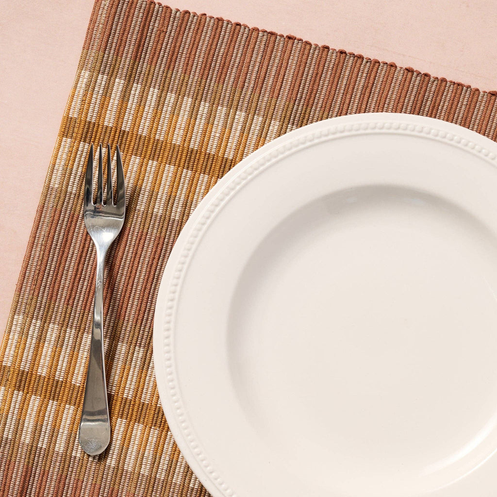 Lifestyle — white plate and fork on placemat (blush bg): "Handloomed brown striped cotton placemat styled with a white dinner plate and silver fork on a blush background"