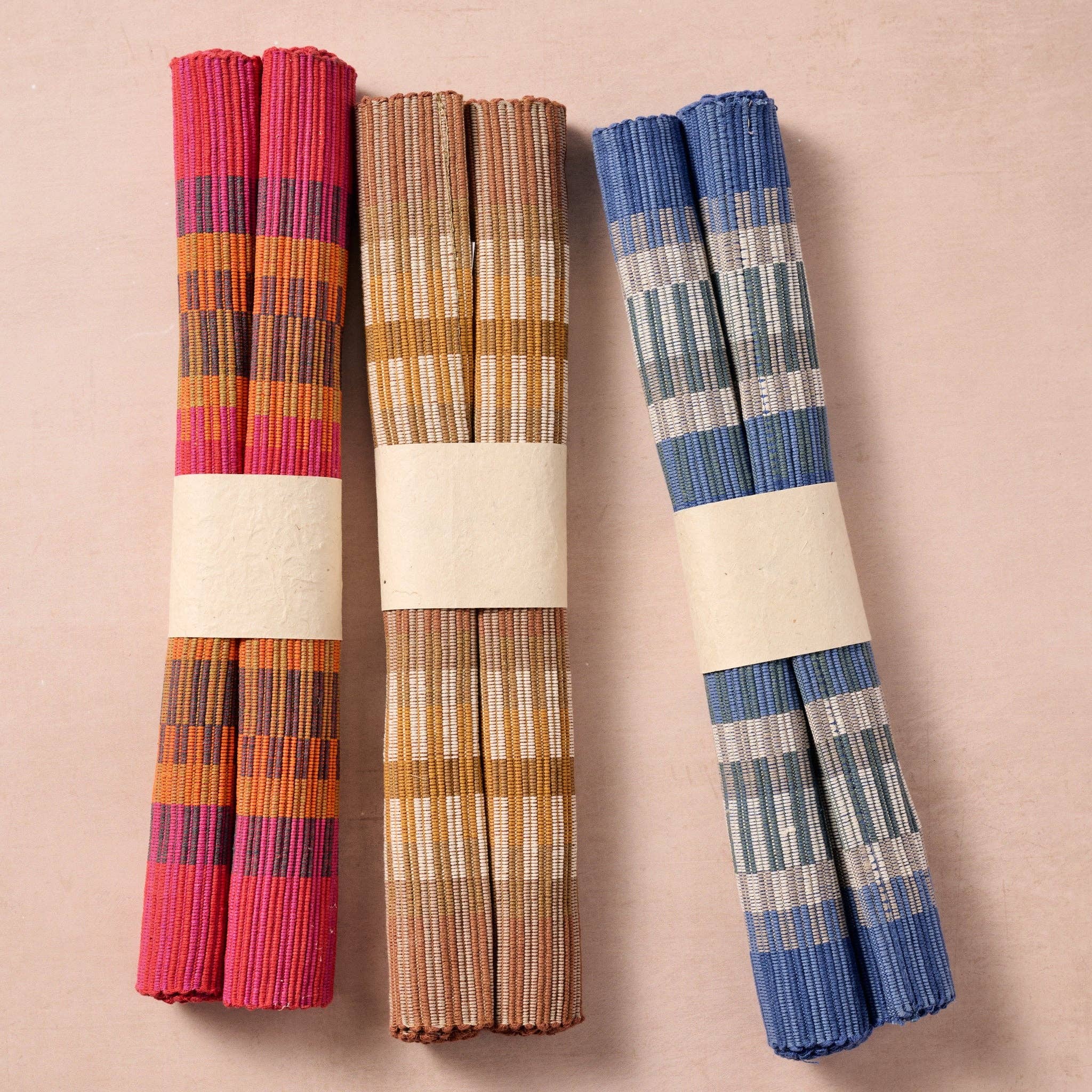 Three colorways rolled side by side: "Three colorways of handloomed cotton placemats — pink, tan, and blue — rolled and displayed side by side"