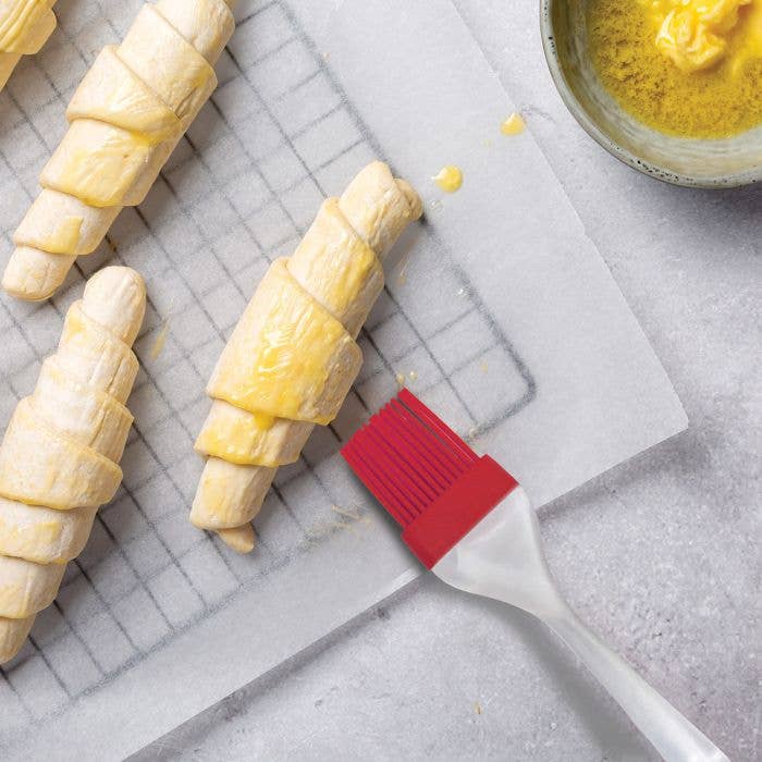 Close-up of Mrs. Anderson's red silicone pastry brush applying egg wash to unbaked croissant rolls on a wire rack, with a small bowl of egg wash nearby.