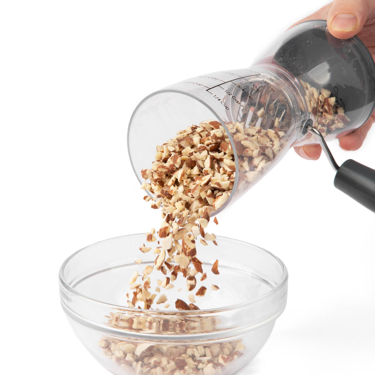 Hand tilting the Progressive nut chopper to pour finely chopped nuts into a clear glass mixing bowl, showing the easy-pour design and fine chop results.