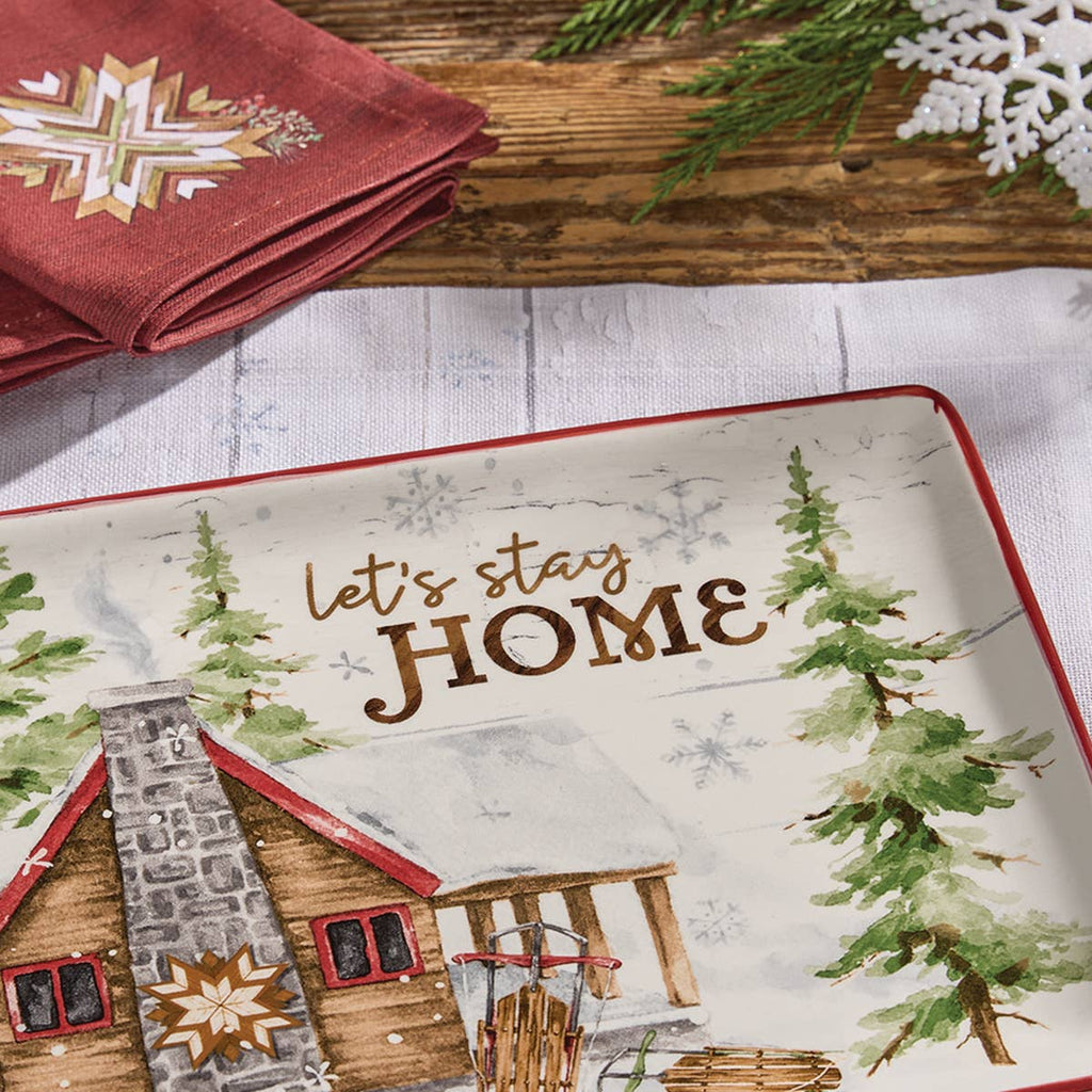 Angled close-up of Park Designs Snowflake Lodge platter on a white linen runner with red napkin and pine branches, showing Let's Stay Home detail"