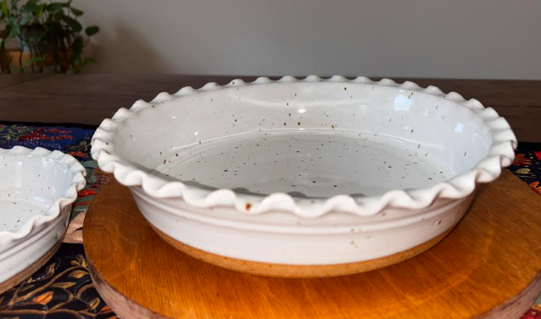 9" angled, both dishes visible, colorful textile) Angled view of two hand-thrown white speckled stoneware deep dish pie plates with ruffled edges on a colorful floral textile