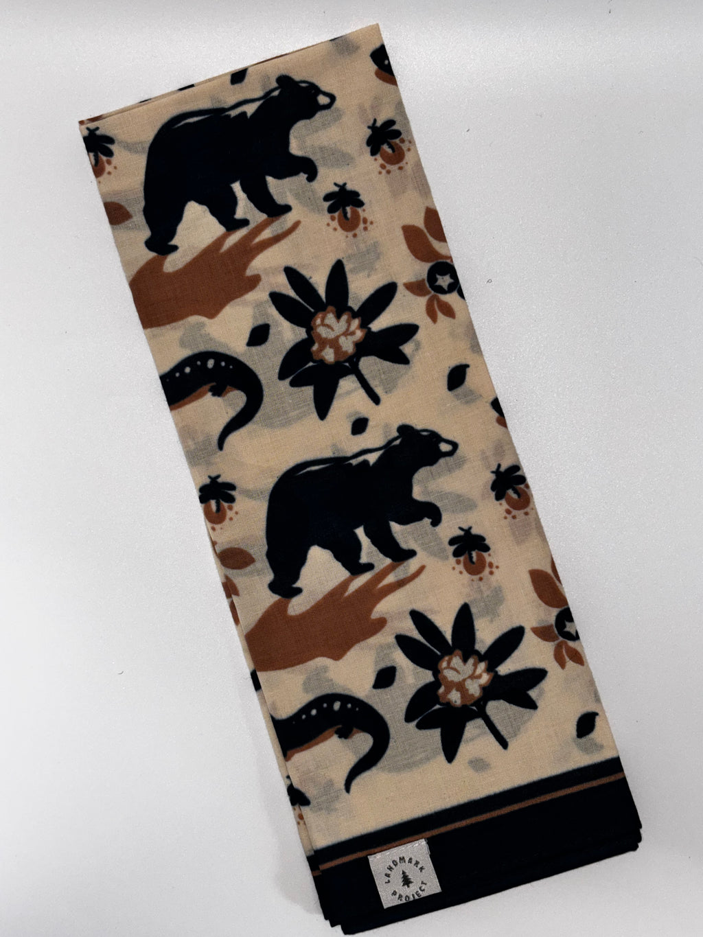 A folded beige scarf featuring a pattern of black bears, brown flowers, and small red ladybugs, with a black border and a small white brand tag at the bottom.