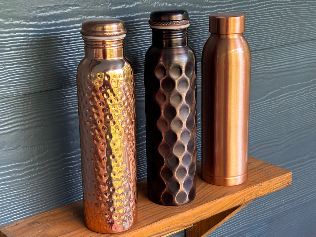 Three Perilla Home pure copper water bottles 1L — Hammered, Antique Diamond, and Cleo styles displayed together on a wooden shelf against a blue-gray siding backdrop