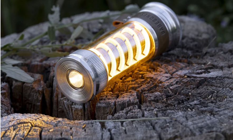 Barebones Edison Light Stick glowing in lantern mode resting on a weathered wood stump outdoors — warm amber LED light with flashlight beam visible"