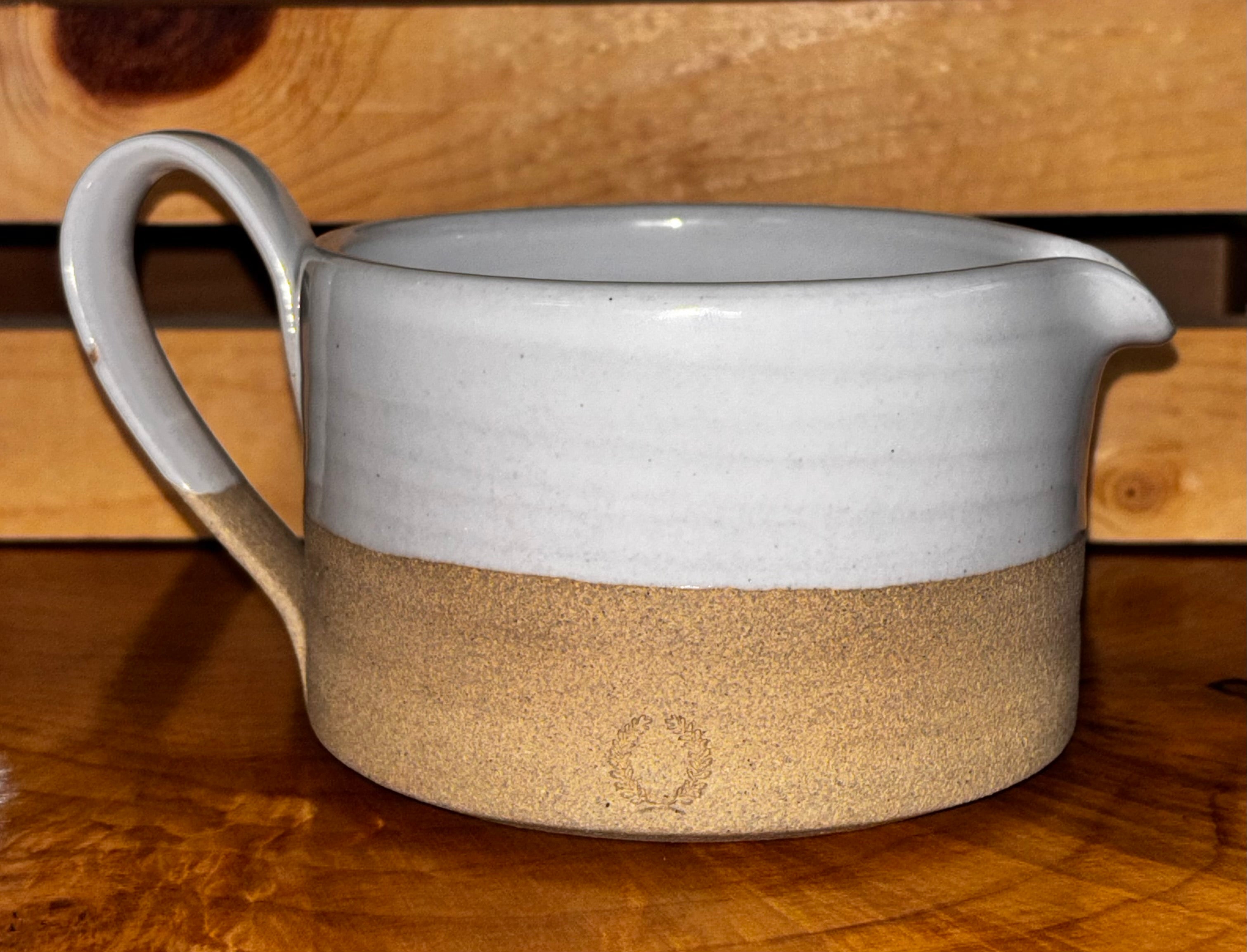 Front view on live edge wood, pallet background → "Farmhouse Pottery Silo stoneware gravy boat with white glaze top and natural unglazed bottom, displayed on a live edge wood surface with rustic pallet background showing the Farmhouse Pottery impressed mark"