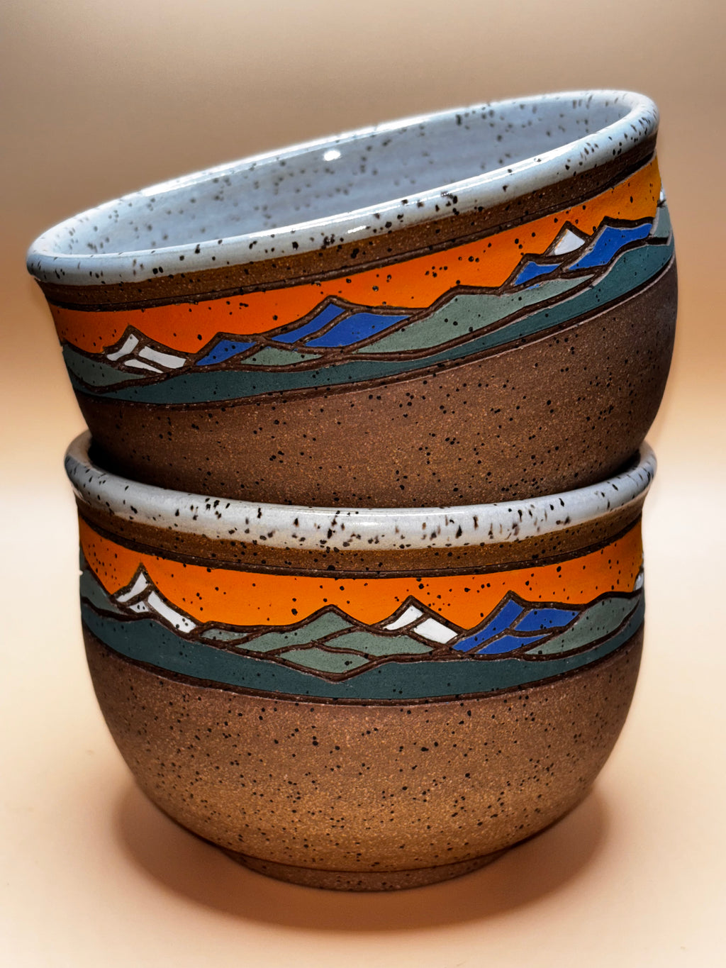Sunset Peak (stacked, tilted interior view):

"Set of 2 handmade speckled stoneware bowls with vibrant orange sunset and blue mountain landscape band, stacked showing speckled white interior — Forest Jeannie Pottery"