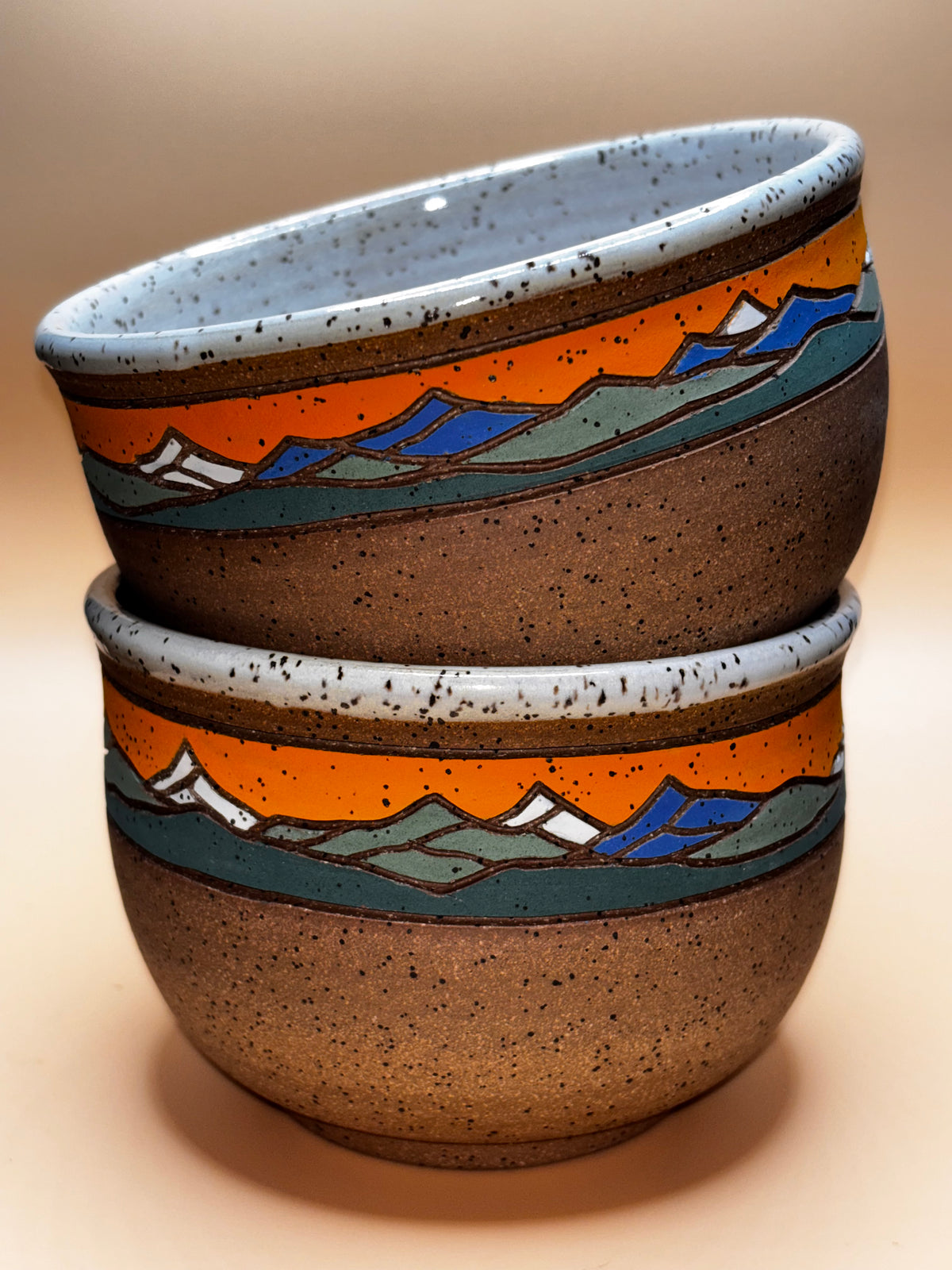 Sunset Peak (stacked, tilted interior view):

"Set of 2 handmade speckled stoneware bowls with vibrant orange sunset and blue mountain landscape band, stacked showing speckled white interior — Forest Jeannie Pottery"