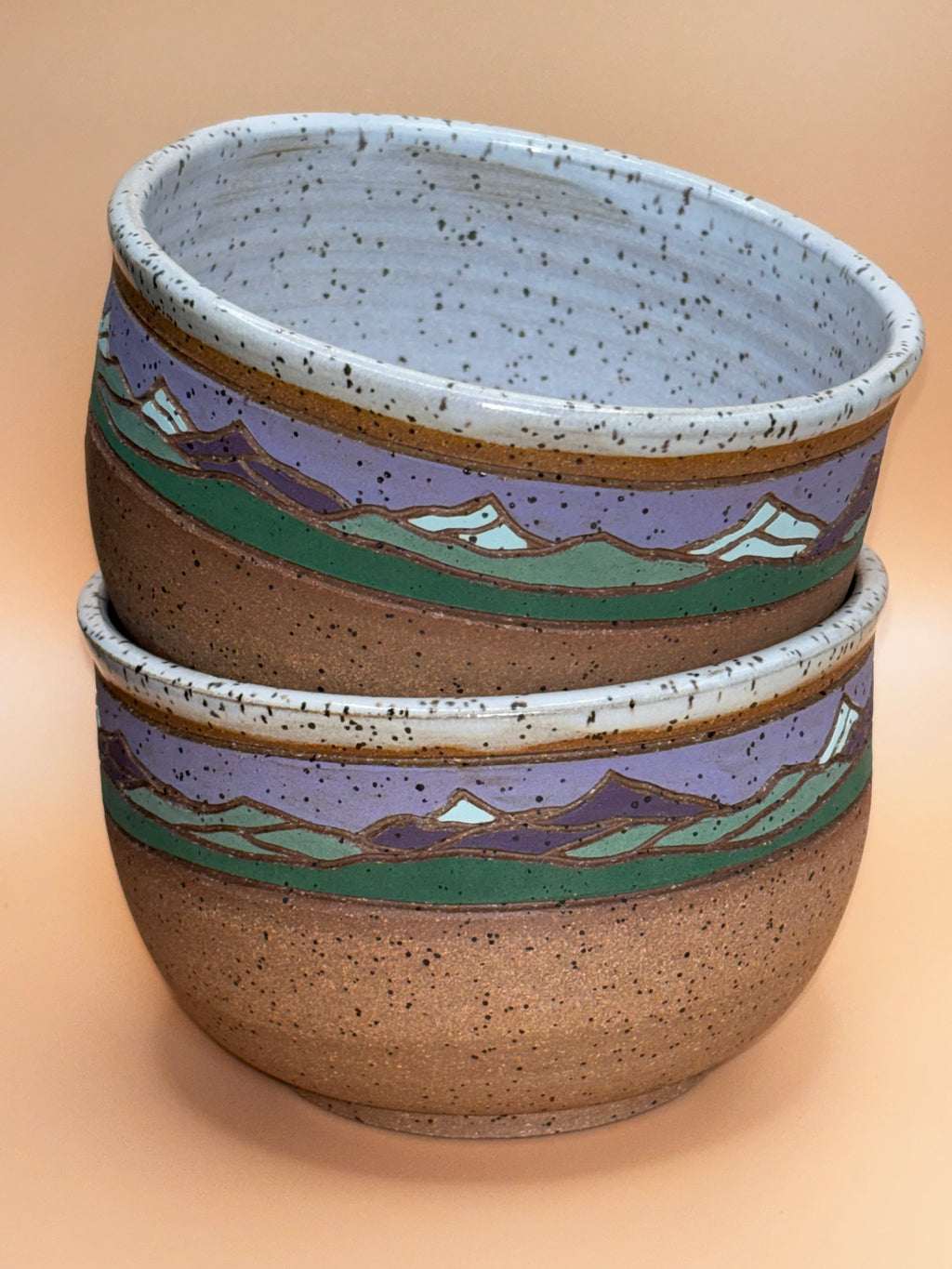 Twilight Summit (stacked interior view):

"Set of 2 handmade speckled stoneware bowls with soft lavender and sage green mountain landscape band, stacked showing speckled white interior — Forest Jeannie Pottery"
