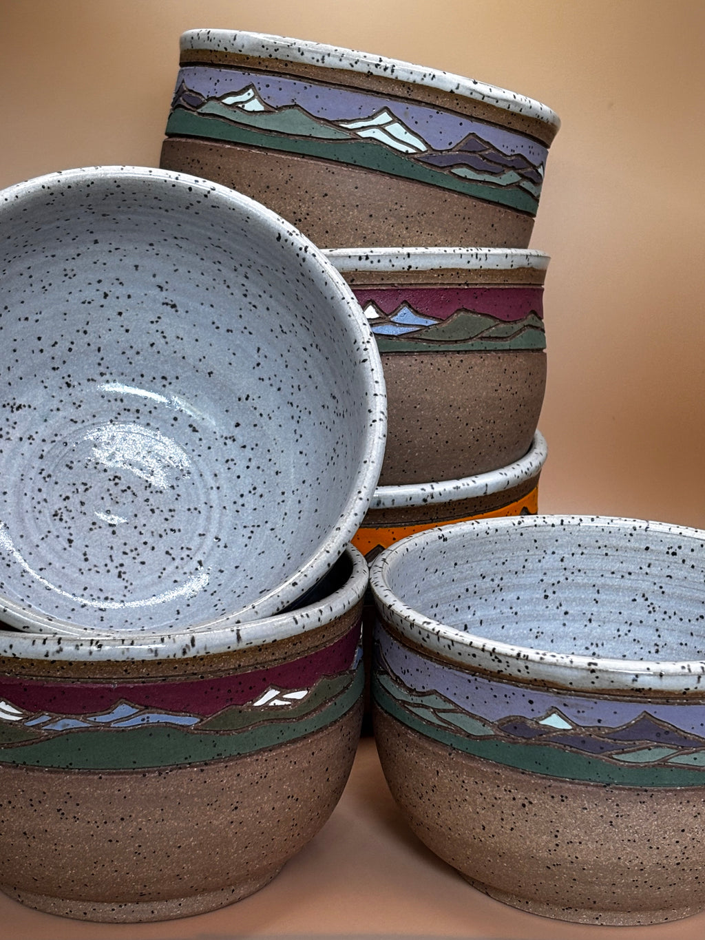 Close-up of all six Forest Jeannie handmade mountain landscape stoneware bowls grouped together, showing speckled white interior and all three colorways — Crimson Ridge, Sunset Peak, and Twilight Summit"