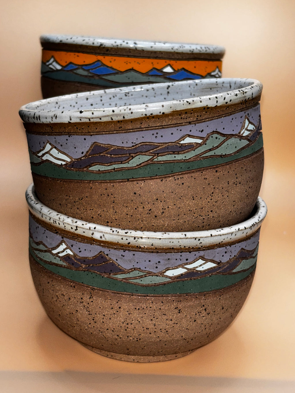 Twilight Summit & Sunset Peak stacked (angled, showing both colorways):

"Two Forest Jeannie handmade stoneware bowl sets stacked — Twilight Summit in soft lavender and Sunset Peak in vibrant orange, showing mountain landscape detail"