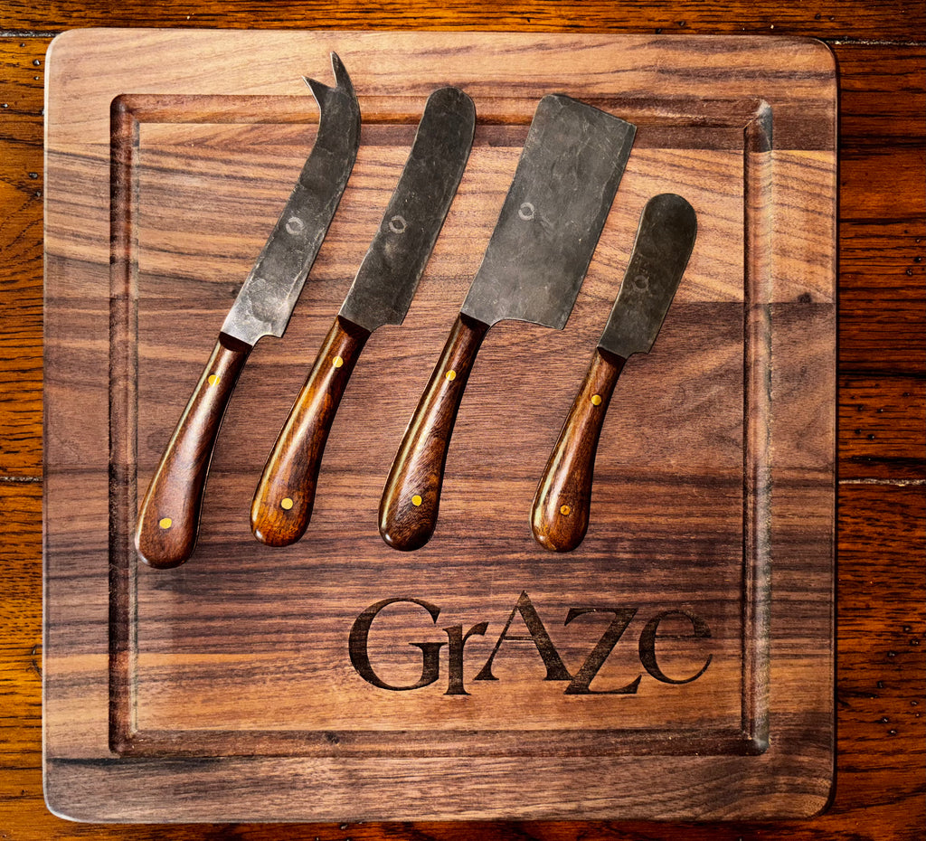 "Set of four Farmhouse Pottery artisan forged cheese knives displayed on an engraved walnut GrAze charcuterie board, showing the hammered steel blades with the Farmhouse Pottery maker's mark and walnut handles with brass rivets, overhead view"