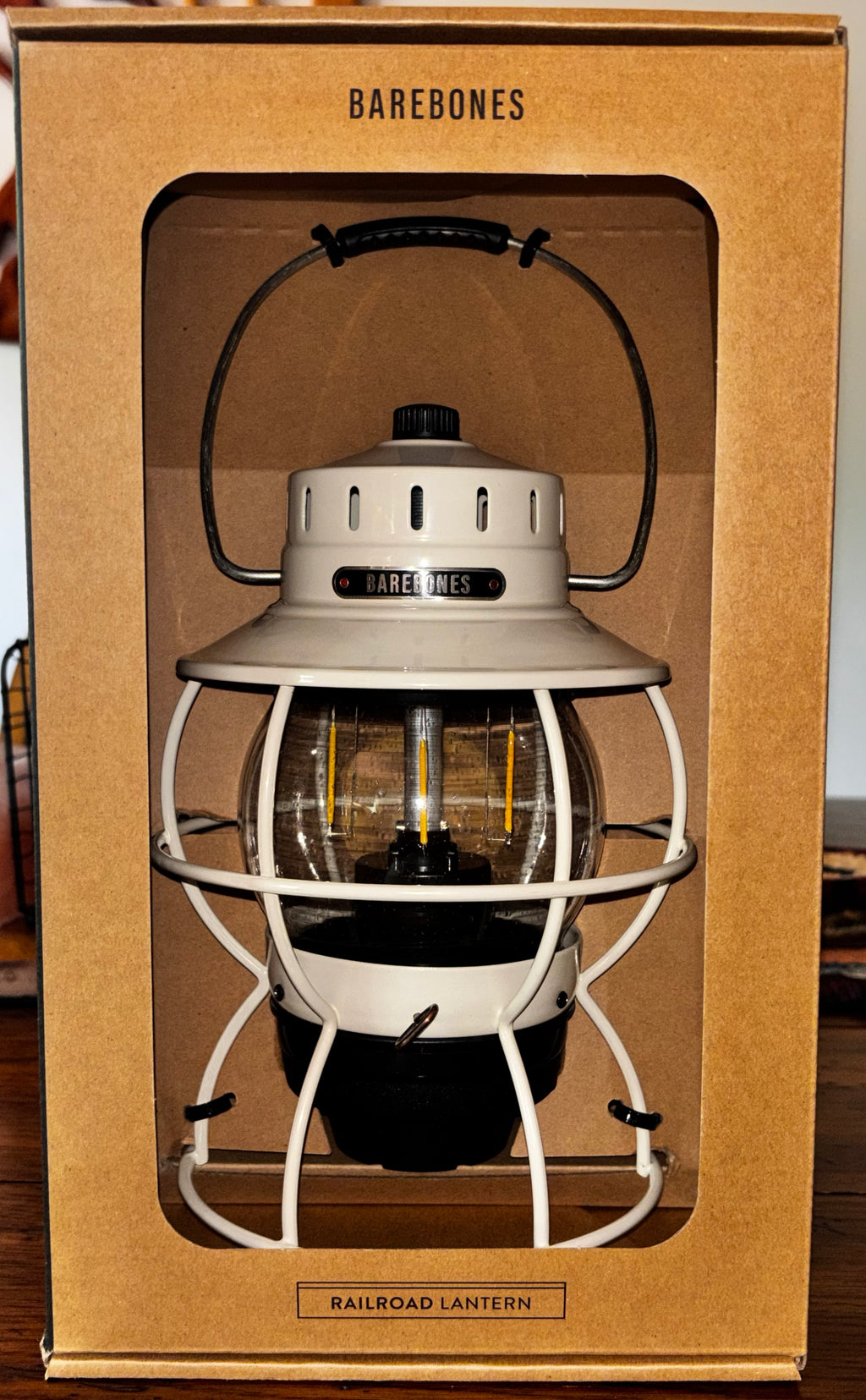 A Barebones Railroad Lantern displayed inside its original brown cardboard packaging box, featuring a cream-colored metal body, black accents, and a clear glass lantern section.