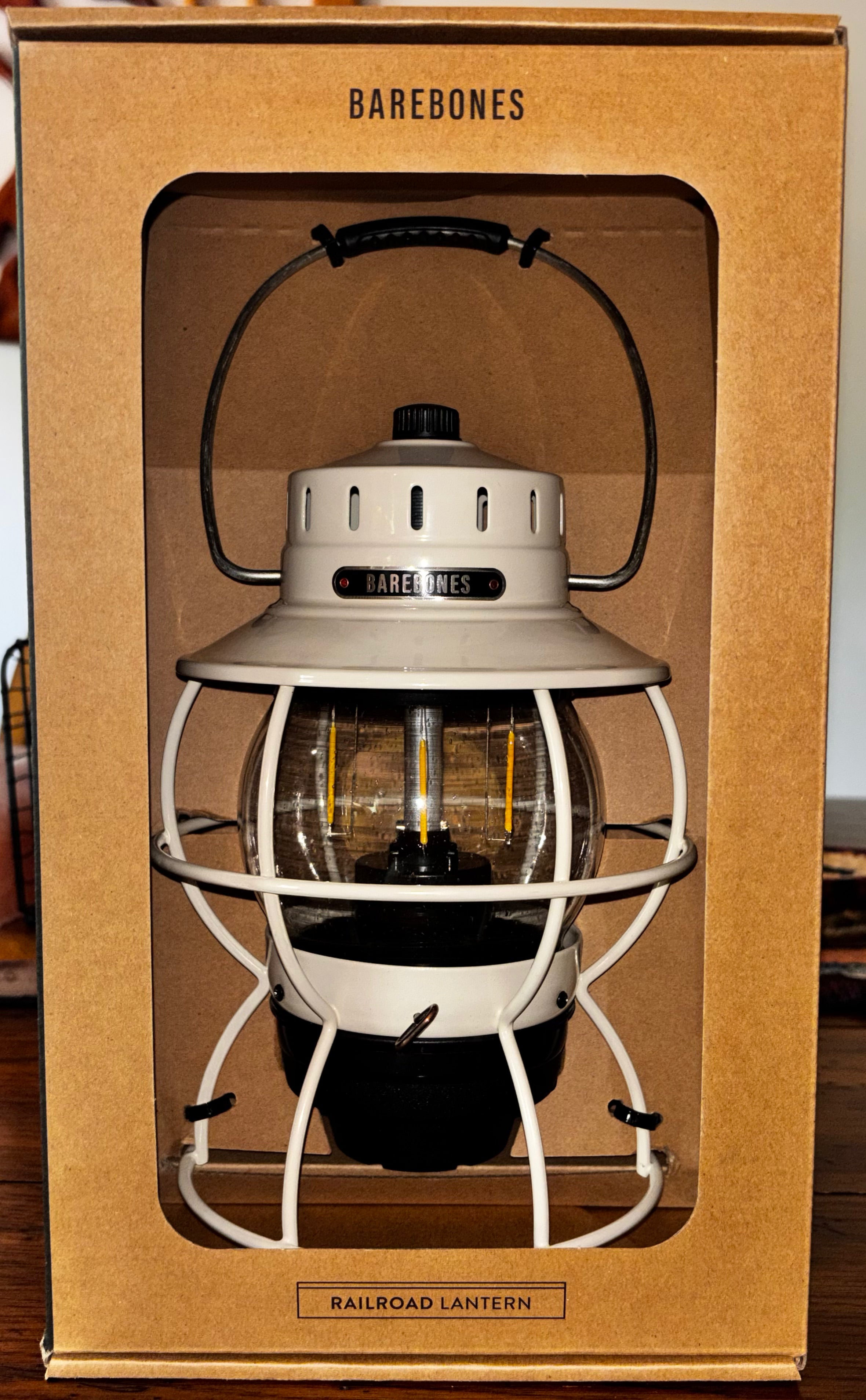 A Barebones Railroad Lantern displayed inside its original brown cardboard packaging box, featuring a cream-colored metal body, black accents, and a clear glass lantern section.