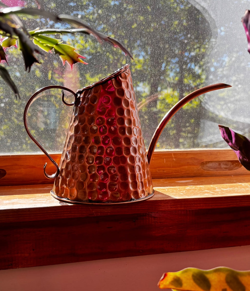 A hammered copper watering can with a long spout and curved handle, resting on a wooden windowsill with natural light shining through the window behind it.