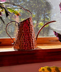 A hammered copper watering can with a long spout and curved handle, resting on a wooden windowsill with natural light shining through the window behind it.