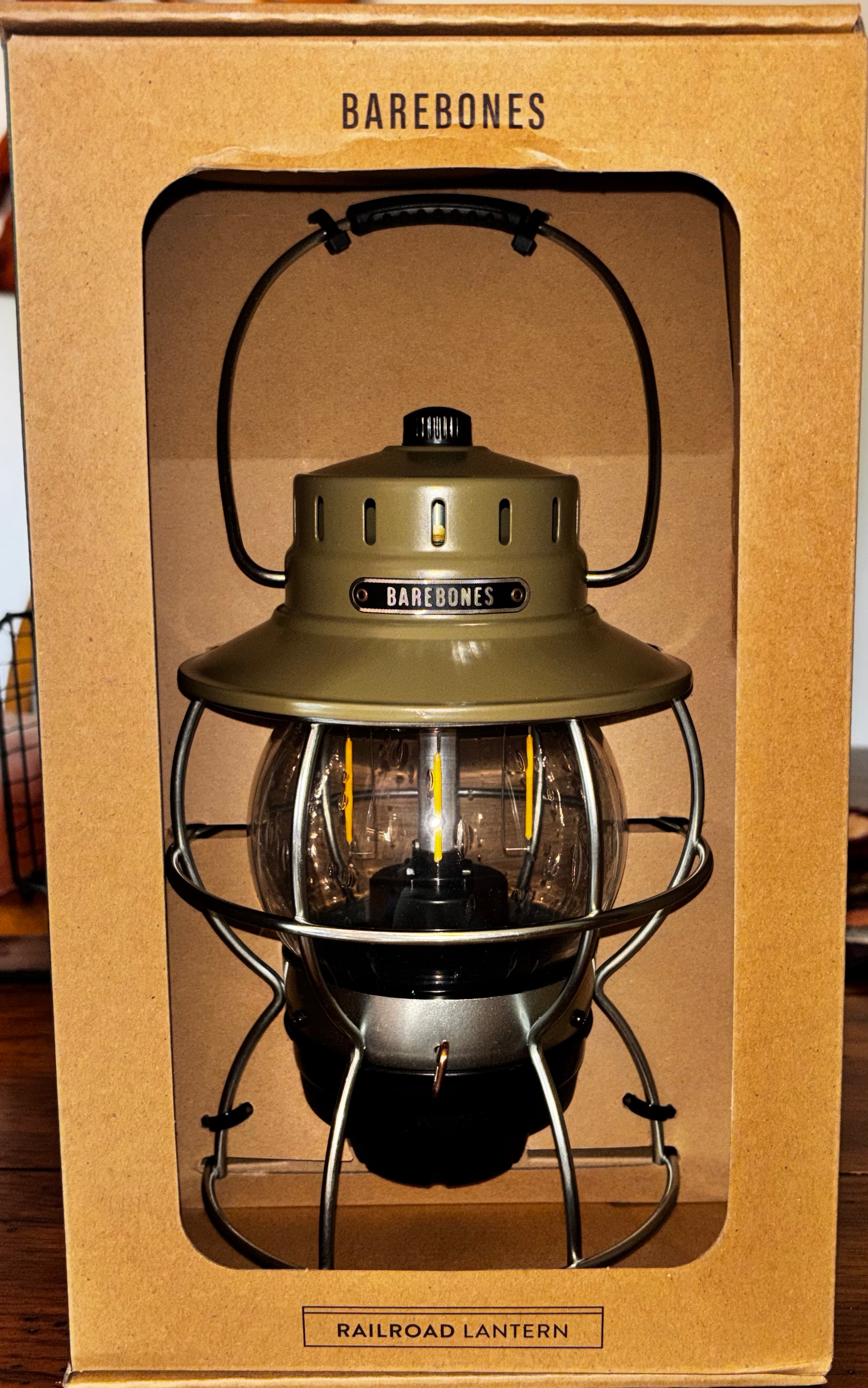 A Barebones Railroad Lantern in its original brown cardboard packaging, featuring an olive green metal body, black wire cage, and clear glass globe.