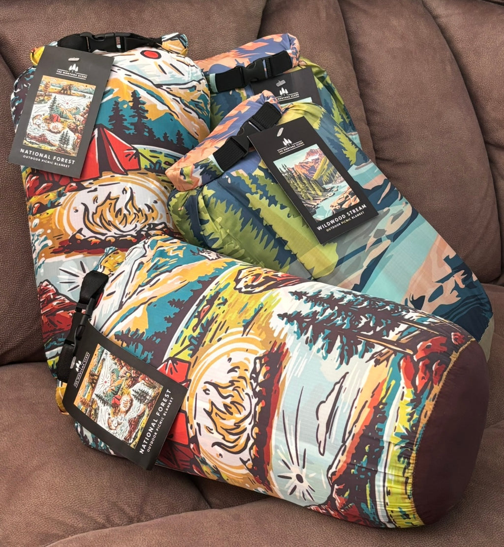 The Montana Scene outdoor picnic blankets rolled and secured in carrying bags with original tags — National Forest and Wildwood Stream designs shown together on a sofa"