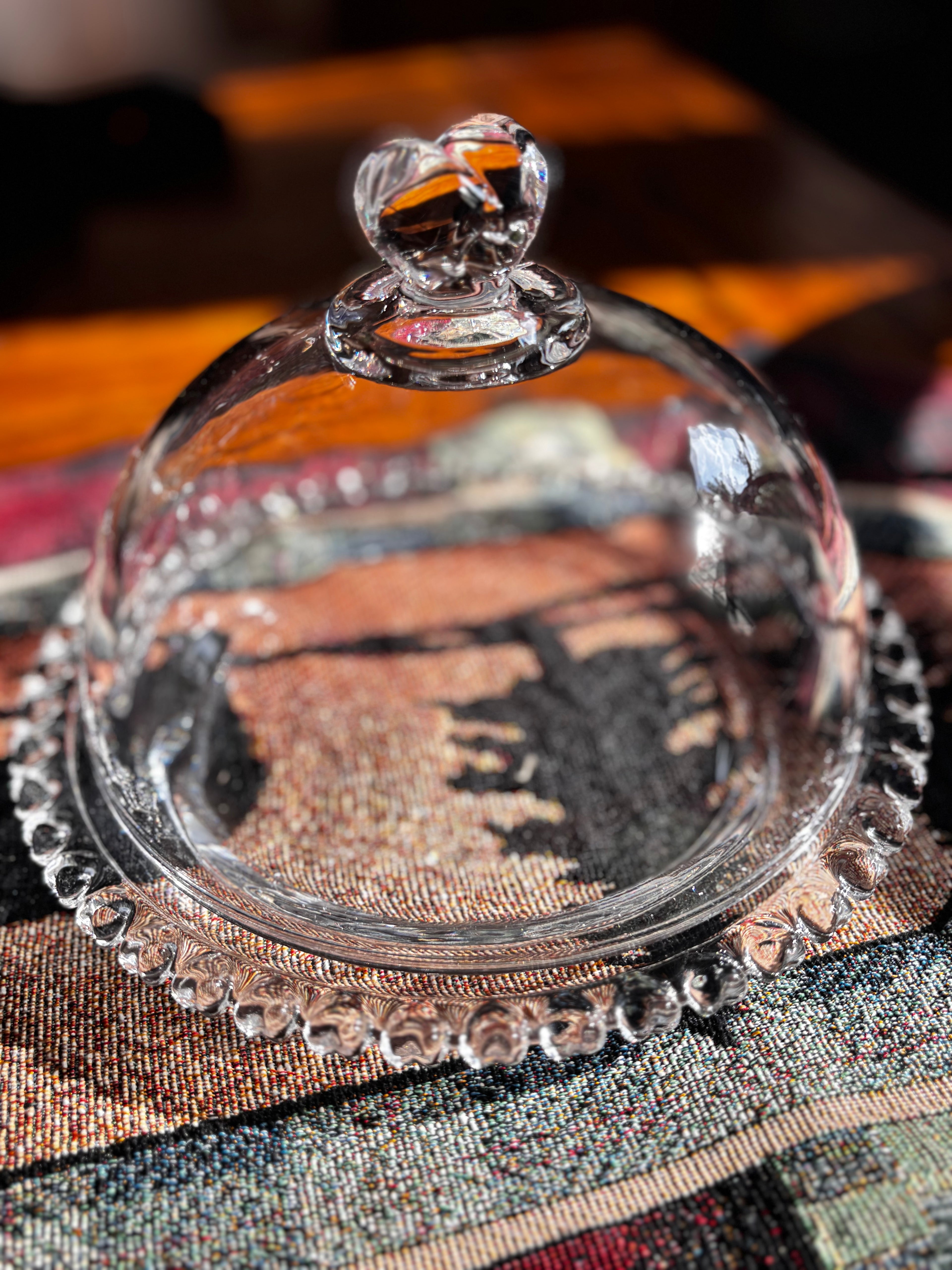 Clear glass cloche with a heart-shaped lid knob and a scalloped rim, photographed close up on a patterned textile surface.